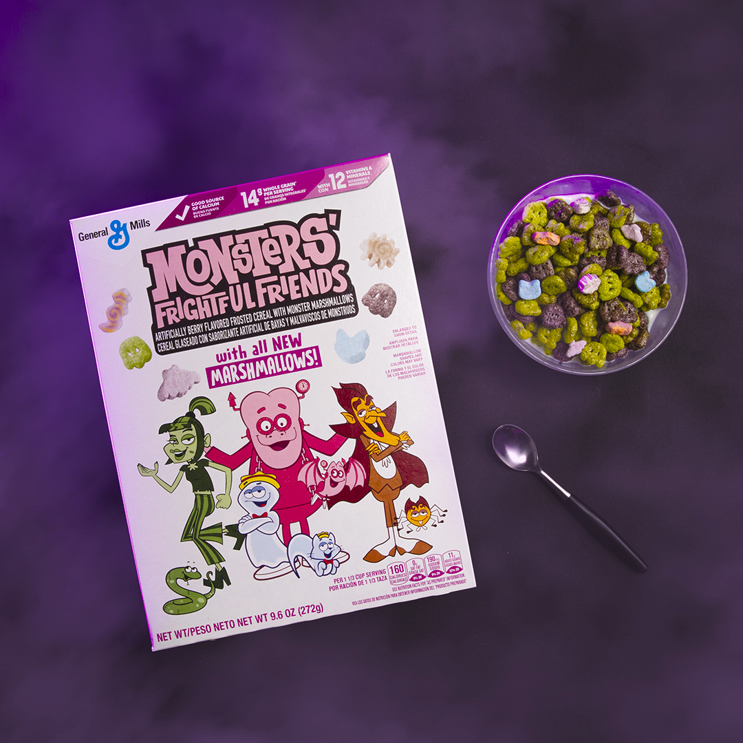 A box of General Mills' Monsters’ Frightful Friends Cereal alongside a bowl filled with colorful cereal and pet-shaped marshmallows, set on a moody purple background with a spoon to the side.